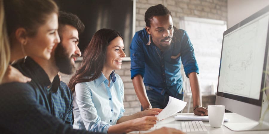 Four smiling colleagues are gathered at a desk, look at a computer screen.
