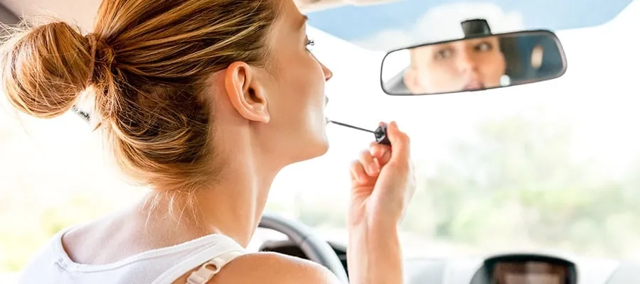Woman applying lip gloss in the car while looking in rearview mirror.